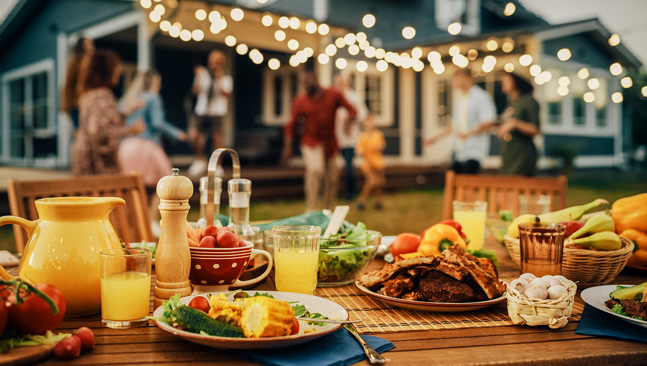 Table of outdoor barbeque food with guests dancing in the background.