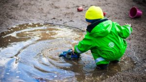 Child in bright yellow hat and green rain suit playing with a toy car in a muddy puddle.