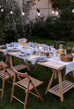 Outdoor dining setup with a table, chairs, and hanging lights in a garden at dusk.