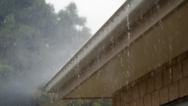 Rain pouring down from the edge of a house roof against a blurred tree background.