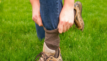 A person adjusts their sock over a pant leg while kneeling on grass.