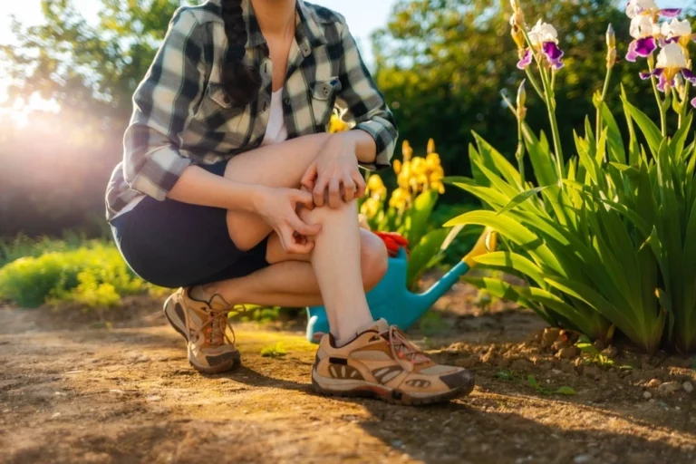 A woman scratching a mosquito bite while watering flowers outside.