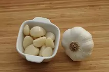 Small, square, ceramic bowl filled with peeled garlic cloves next to a whole garlic bulb on top of a table. 